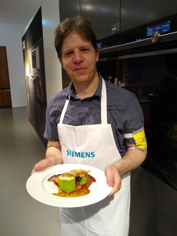 Foto von Thomas Posch mit Kochschürze und lächelnd, mit einem Teller in der Hand, auf dem selbstgekochtes Abendessen präsentiert wird